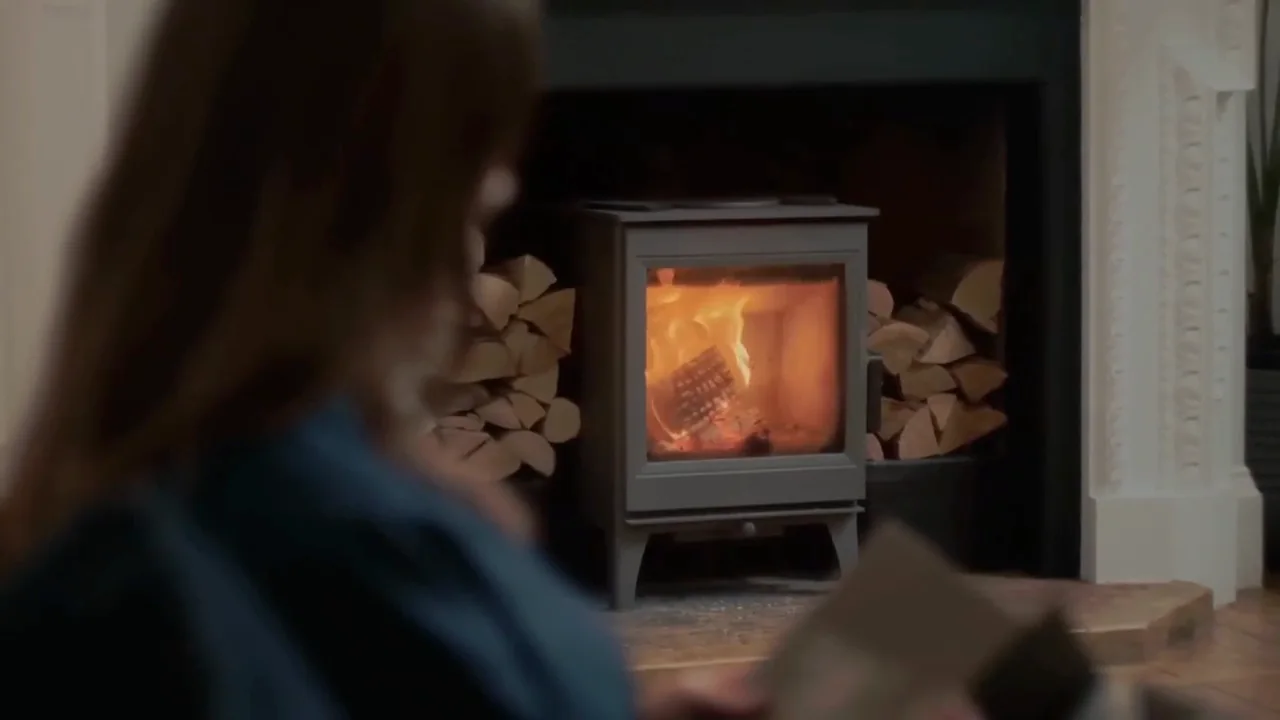 A woman reading in the foreground while a wood-burning stove burns behind her, logs stacked either side