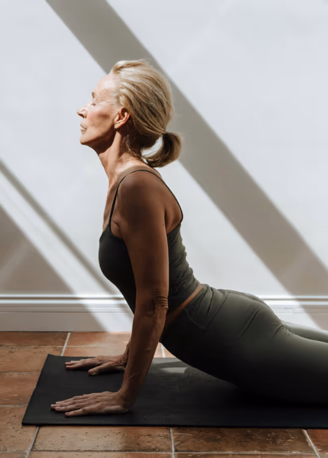 Older woman with a blonde ponytail in an upward-facing yoga pose on a mat, with striped sunlight on the wall behind her