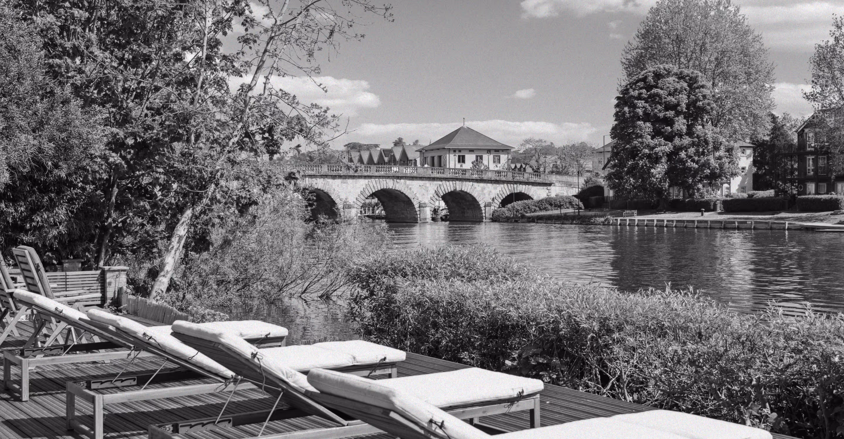 Black-and-white view of sun loungers on a riverside deck looking across the Thames to a stone bridge