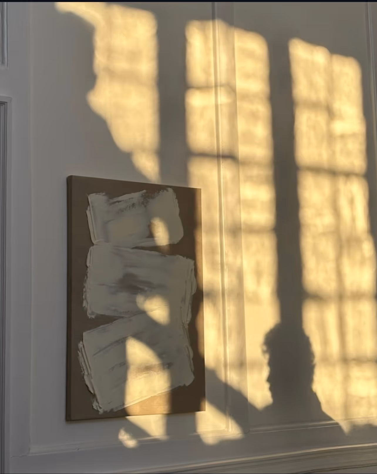 A textured abstract canvas on a wall with golden window-grid shadows and a person's silhouette cast across it